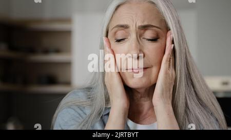 Sick old mature caucasian woman worried tired upset retiree grey-haired lady holding head massaging temples unbearable headache pressure suffering Stock Photo