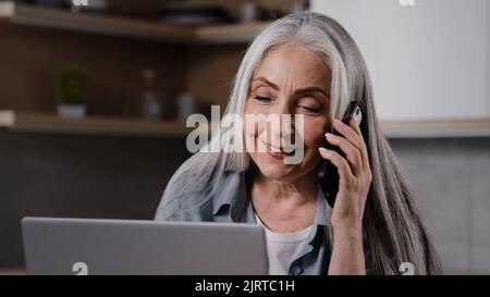 Aged business lady experienced female specialist talk mobile phone look at laptop screen negotiate with client in home kitchen. Grey-haired housewife Stock Photo