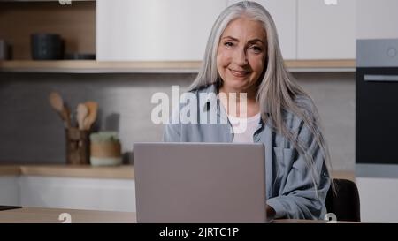 Mature trendy caucasian old businesswoman domestic teacher elderly housewife granny typing on keyboard laptop in kitchen room use home internet check Stock Photo