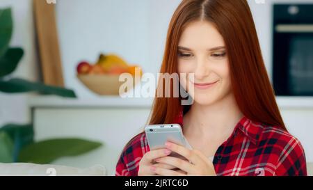 Smiling red-haired caucasian girl model woman sits in kitchen room holds modern gadget smartphone looks at mobile display reads sms messages from Stock Photo
