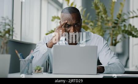 Concentrated african man employee american expert focused specialist sits in office works on laptop use personal computer typing email chatting Stock Photo