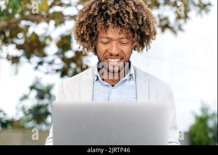Happy successful mixed race curly haired male top manager, freelance, ceo, stylishly elegantly dressed, male business person, sitting outdoors, working in a laptop on a project, smiling friendly Stock Photo