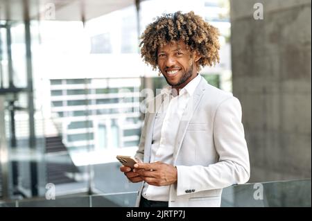 Handsome happy brazilian or hispanic man with curly hair, stylishly elegantly dressed, standing outdoors near the business center, using mobile phone, online messaging, looks at camera, smiling Stock Photo