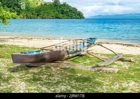 An old outrigger boat used to navigate from island to island in Fiji sits on the sandy shoreline framed by a beautiful blue sky and ocean. Stock Photo