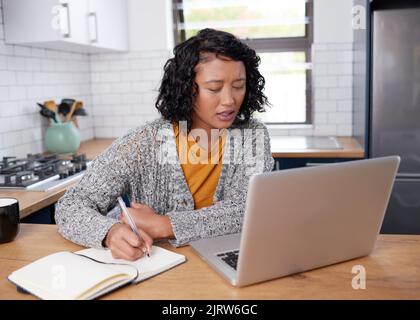A young multi-ethnic woman works from home in the kitchen taking notes Stock Photo