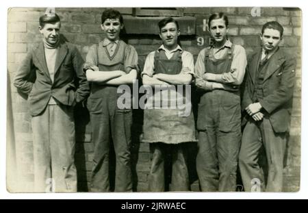 Original 1930's era postcard of five cheerful looking working class lads, possibly on apprenticeships, in industry or construction. One young man is wearing an apron, 2 are wearing overalls, 2 are wearing suit jackets and trousers, unknown location in the U.K. Stock Photo