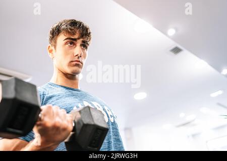 From below determined young sportsman doing bicep curl exercise with heavy dumbbell and looking away against white ceiling in modern gym Stock Photo