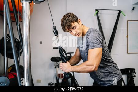 Side view of strong male athlete pulling rope and checking bicep while doing exercise with cable machine in modern gym Stock Photo