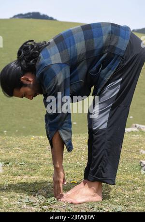 Side view of a long haired Indian young guy doing Uttanasana Yoga Pose in the mountain. A male hiker doing stretching by Standing Forward Bend Toe Touch exercise. Stock Photo