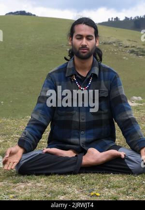 Front view of a good looking Indian young man practicing Yoga in Lotus Pose (Padmasana) with Gyan Mudra (Gesture of Knowledge) in the mountain. Stock Photo