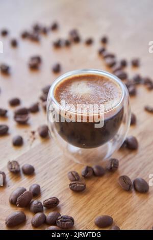 A transparent glass cup of hot espresso standing on a wooden table top, surrounded by coffee beans in the background Stock Photo