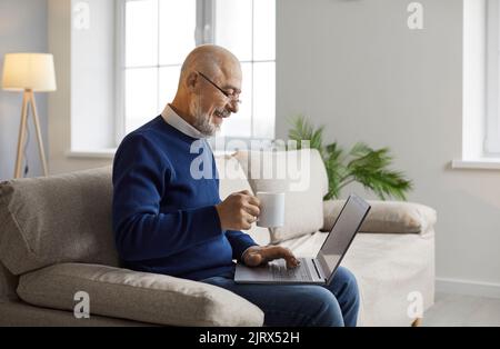 Happy senior man sitting on sofa at home, using laptop computer and drinking coffee Stock Photo