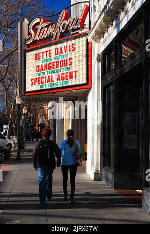 A young adult couple walk while holding hands under a marquee for a movie theater showing classic Hollywood films starring Bette Davis Stock Photo