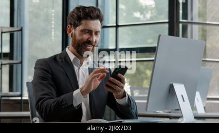 Bearded middle-aged business man in office sitting table using phone mobile audio app service to listening music singing song with modern wireless Stock Photo