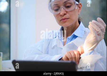 Close-up female scientist in white lab coat and safety goggles works on digital tablet and puts on protective gloves Stock Photo