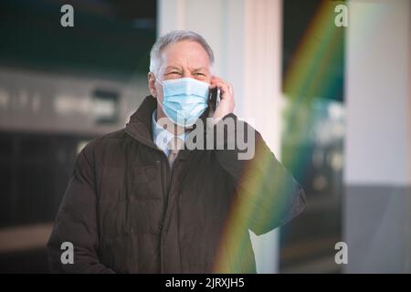 Covid coronavirus concept, masked elder business man talking on his smartphone in a train station. Controlled lens flare Stock Photo