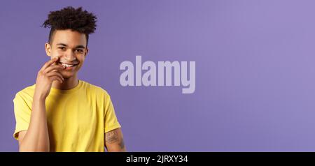 Close-up portrait of lovely young hispanic gay man with dreads, tattoos, touching lip sensually and flirty smiling, checking out someone really cute Stock Photo