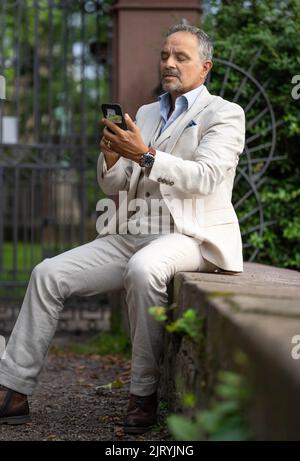 Man in suit sitting on a wall and looking strained into his phone, Karlsruhe, Germany Stock Photo