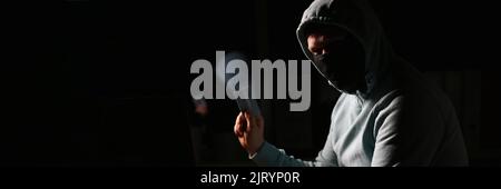 Hacker in dark jacket looking at laptop screen in dark room is holding bank card Stock Photo