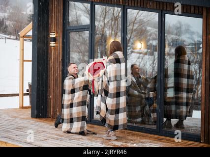 Happy man in love making marriage proposal to girlfriend under winter snow while woman holding bouquet of flowers. Couple sharing romantic moment outside scandinavian house barnhouse. Stock Photo