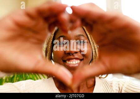 hispanic woman showing love with hands in heart shape expressing healthy and marriage symbol near window and green plant at home Stock Photo