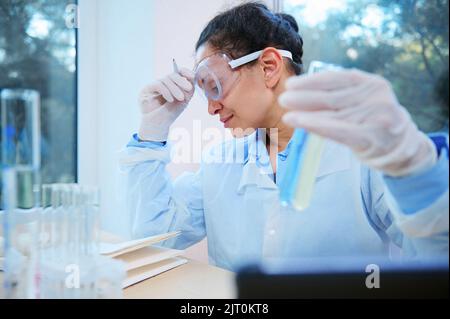 Female scientist pharmacologist holds test tube with blue liquid substance, doing medical research, works in laboratory. Stock Photo