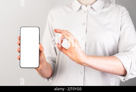 Woman holding phone mockup and showing ok gesture. Female approving, recommending mobile app to clients. Smartphone with white template. High quality Stock Photo