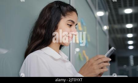 Side view young smiling woman browsing social network makes order in online store using smartphone remote communication in friendly chat on phone Stock Photo