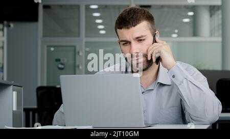 Concentrated young businessman speaks on phone consults client checks data on laptop screen serious manager remote seller offers services over Stock Photo