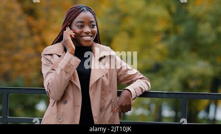 Young confident woman standing outdoors talking on telephone answering business call remotely orders service using mobile phone chatting with friends Stock Photo