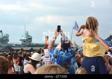 Festival goers at the Victorious Festival on Southsea Common give Royal Navy aircraft carrier HMS Prince of Wales a colourful send-off while listening to the Sugababes as it sails from Portsmouth Naval Base, Hampshire, for exercises with the US Navy off the coast of America. Picture date: Saturday August 27, 2022. Stock Photo