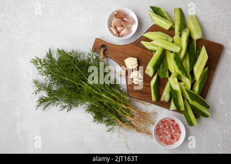 Peeled fresh cucumbers on a brown wooden cutting board with a bunch of dill, garlic and pink salt on a light gray concrete table Stock Photo