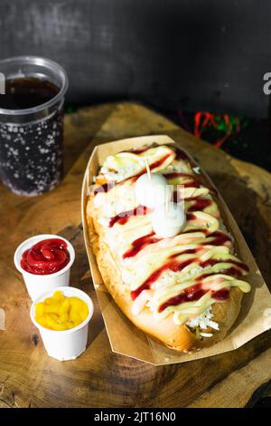 Fast street food, hot dog with sauces, cheese and two quail eggs on top served on a wooden tray and next to it there are two plastic cups with ketchup Stock Photo