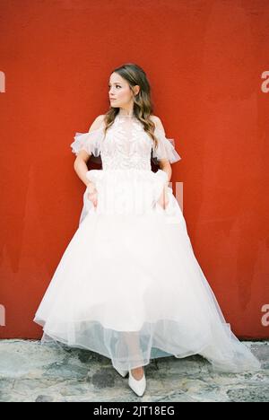 Bride in a white dress stands near a red wall Stock Photo