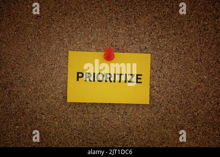 A yellow paper note with the word Prioritize on it pinned to a cork board. Close up. Stock Photo