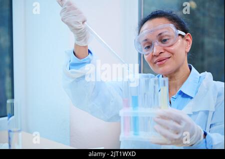 Inspired Hispanic woman lab assistant, scientist chemist using lab dropper dripping few chemical reagent into test tubes Stock Photo