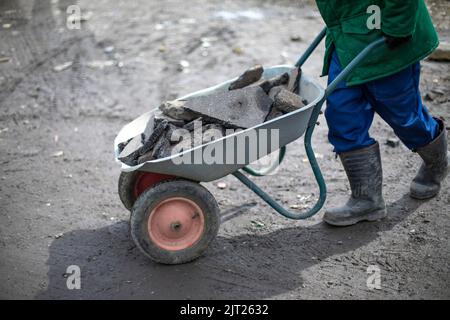 Builder removes broken stone. Man carries cobblestones in cart. Worker in green jacket makes embankment on road. Elimination of pits. Stock Photo