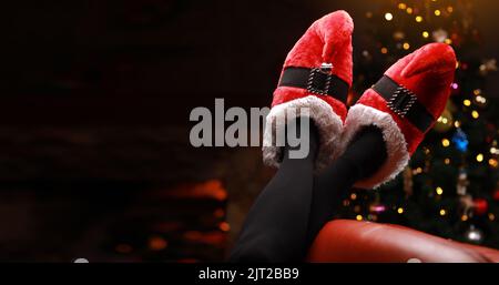 female feet is wearing in Christmas elf shoes on Christmas tree bright background. Woman in elf costume celebration Christmas and New Year at home on couch. Holidays concept. copy space. banner Stock Photo