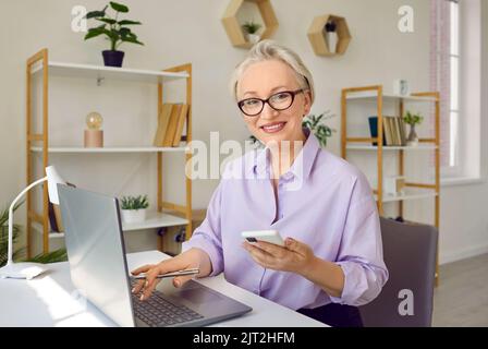 Beautiful smiling senior business lady using mobile phone and laptop in her work. Stock Photo