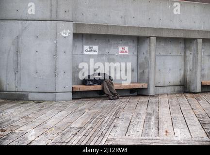Poverty in city man asleep hiding under clothing on bench with signs above banning smoking and camping. Stock Photo