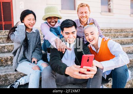 Five young friends looking at mobile phone. Group of people sitting outdoors watching video on smartphone. Stock Photo