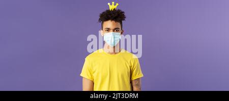 Portrait of funny hispanic guy in face mask, fool around, going crazy staying inside home during quarantine, wearing small crown and look camera Stock Photo