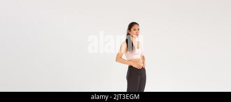 Full length of upset and gloomy asian fitness girl trying to lose weight, touching belly, cant see results from workout or diet, feeling uneasy while Stock Photo