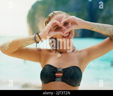 White beaches, warm water - whats not to love. Portrait of a happy young woman making a heart shape with her hands while swimming in the sea. Stock Photo