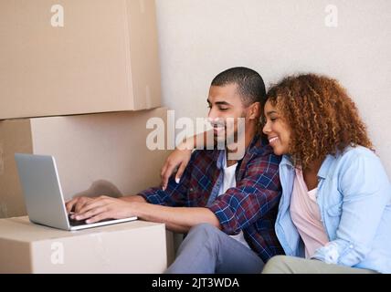 Sending out those housewarming invites. a young couple using a laptop while sitting among boxes on moving day. Stock Photo
