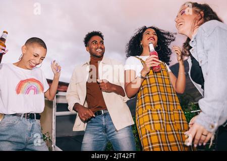 Friends dancing during a rooftop party. Group of happy generation z friends celebrating with beers on a rooftop. Four multicultural young people havin Stock Photo