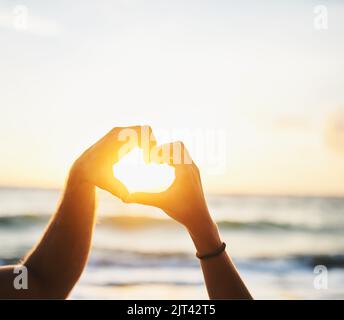 Cherish every sunset. a couple forming a heart shape with their hands at the beach. Stock Photo