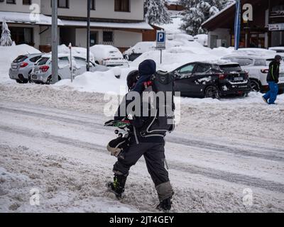 Father Walking down a Snow-covered Street with Helmet and Ski Goggles Carrying his few-month-old Baby on his Shoulders and Holding his Pair of Skis in Stock Photo