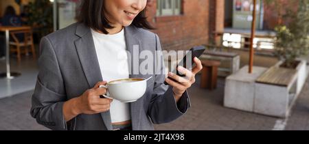 Young elegant professional woman holding coffee standing outside using phone. Stock Photo
