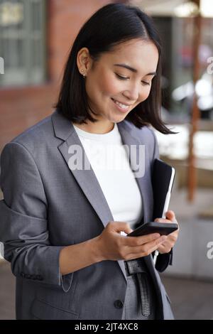 Young happy Asian professional businesswoman wearing suit using phone, vertical. Stock Photo
